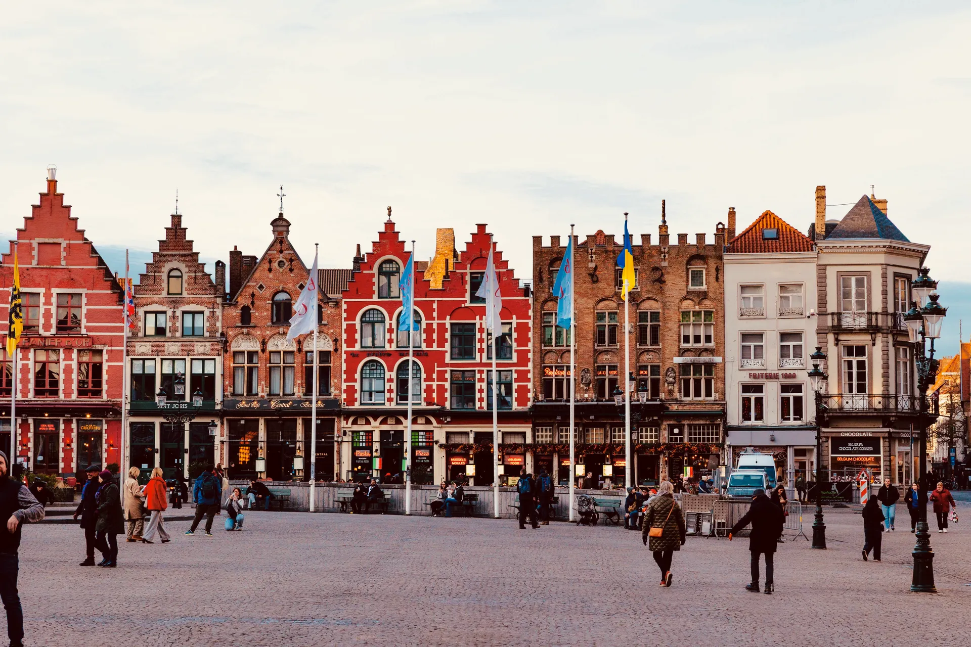 Plaza mayor de una ciudad flamenca al atardecer, con casas de fachadas escalonadas de ladrillo rojo, banderas y gente paseando sobre el adoquín