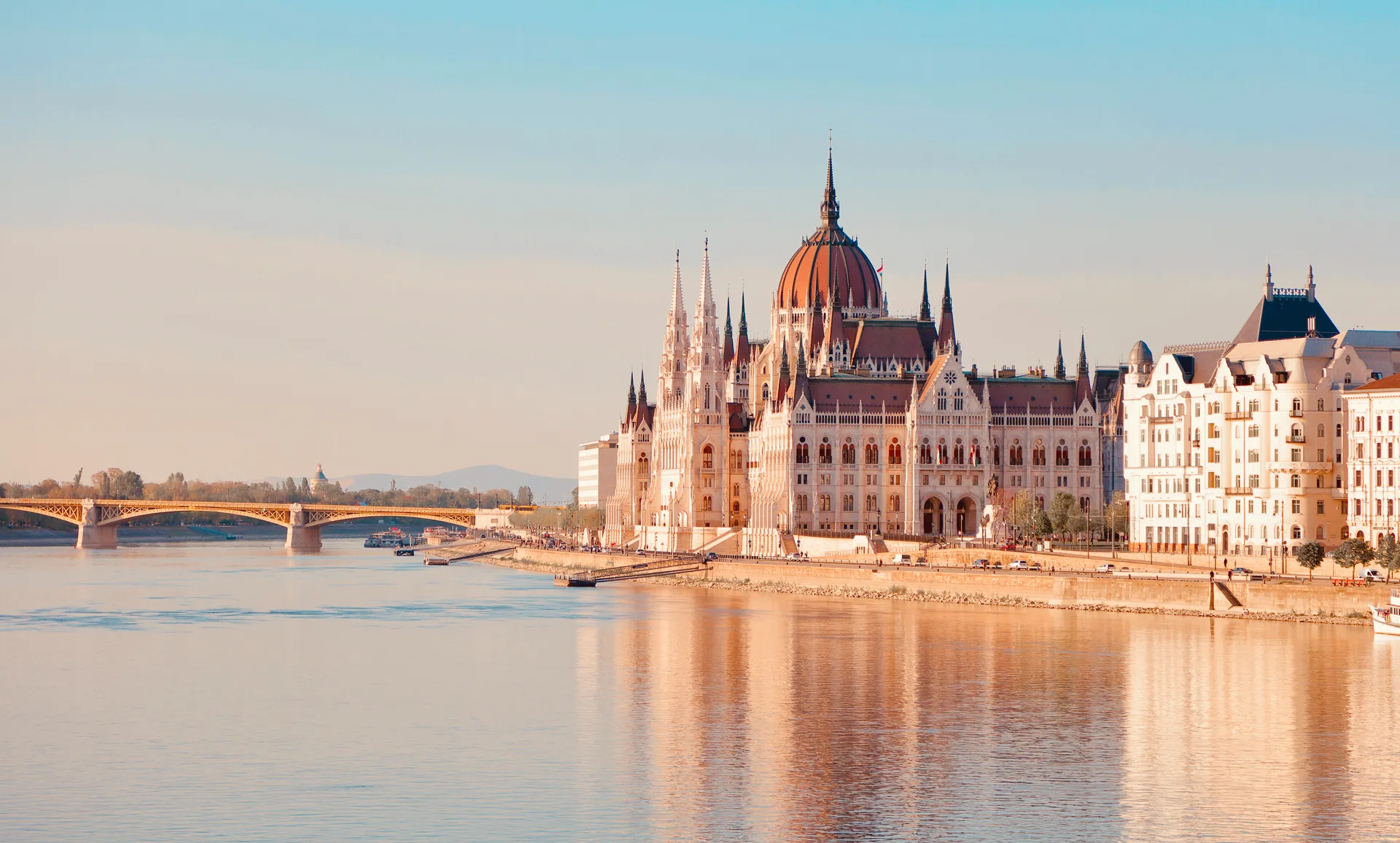 El Parlamento neogótico de Budapest a orillas del Danubio a la luz dorada del atardecer, con un puente al fondo y el edificio reflejado en el agua