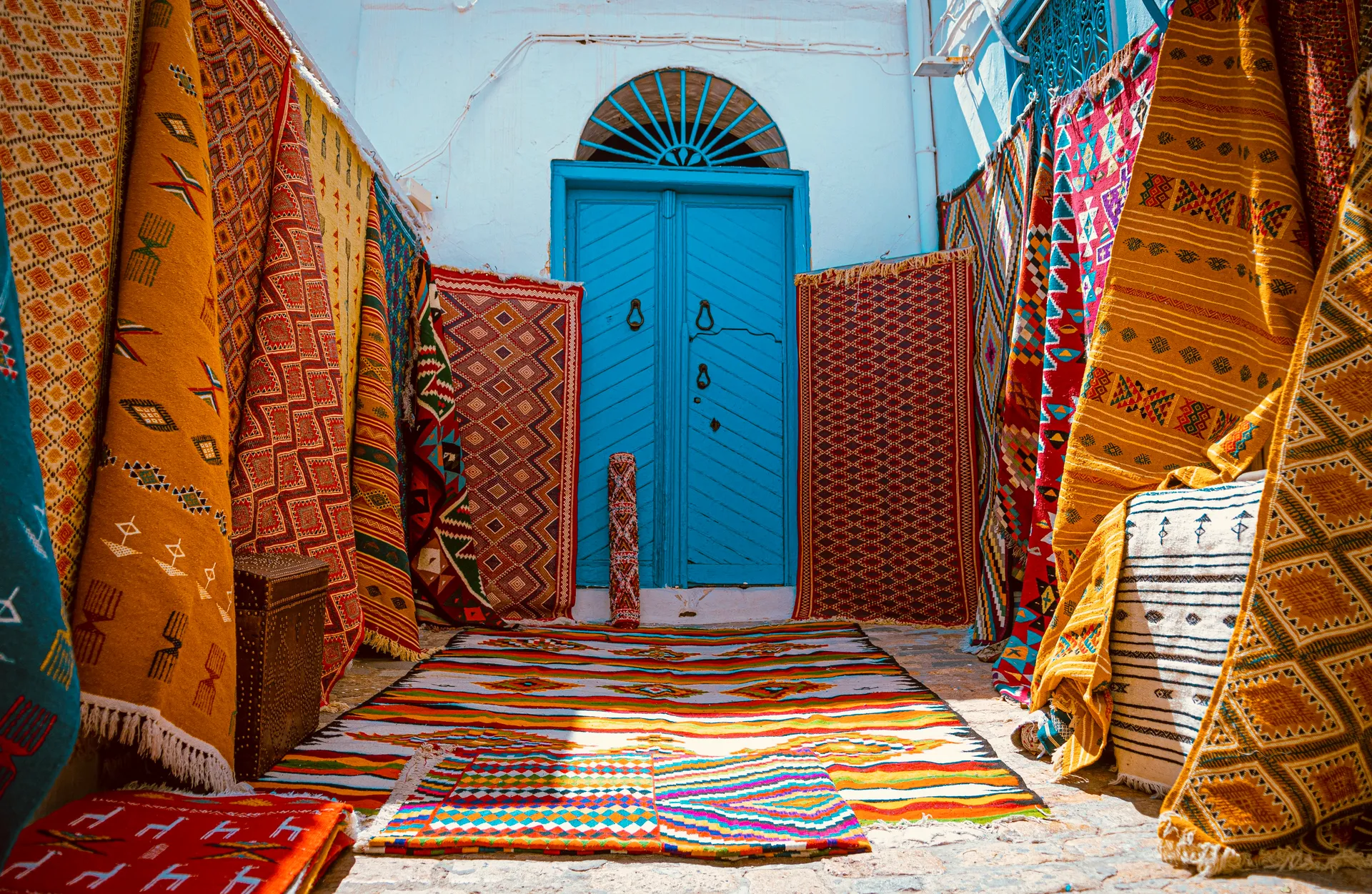 Callejón de un zoco tunecino con alfombras bereberes de colores colgadas a ambos lados y una puerta de madera azul turquesa al fondo, sobre una pared encalada